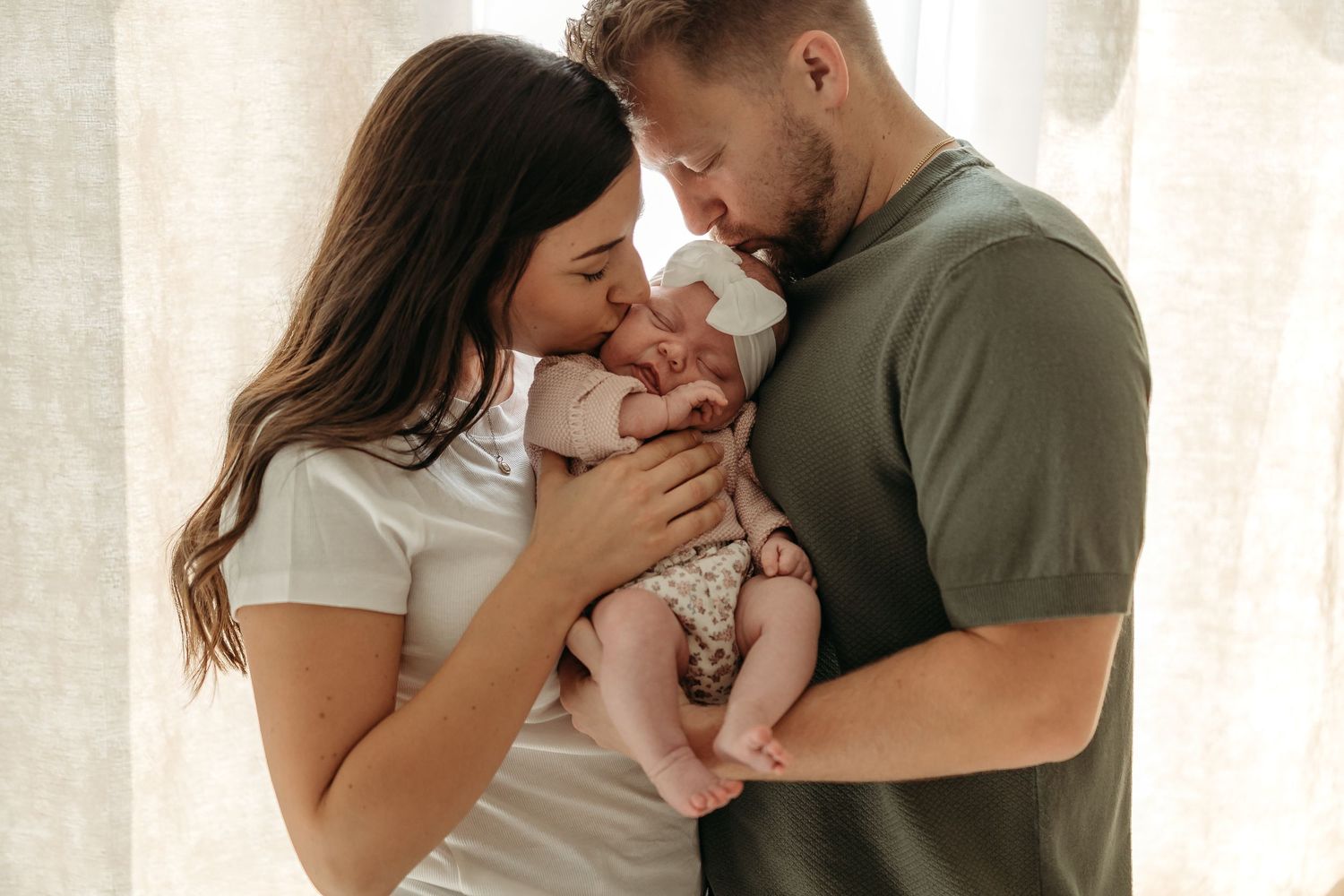 Loving couple in casual sweaters holding small baby while sharing tender moment near window with curtains.
