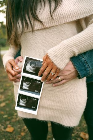 Person in cream sweater holding ultrasound photos outdoors in fall.