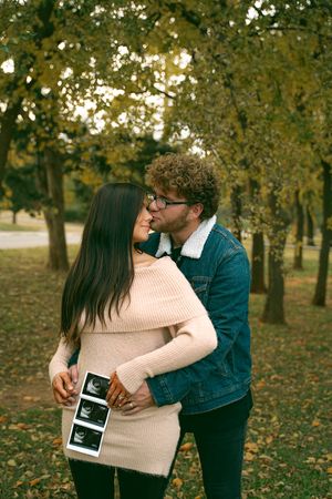 Couple embracing while holding ultrasound photos among fall trees.