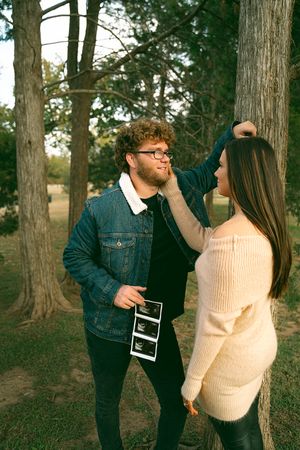 Couple sharing joyful moments with ultrasound photos in wooded park setting.