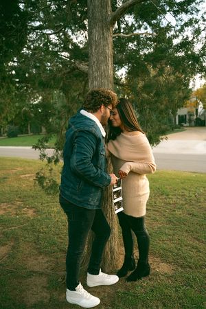 Couple sharing intimate moment by tree during pregnancy announcement photoshoot.