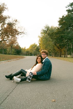 Two people sitting casually on asphalt path surrounded by autumn trees.
