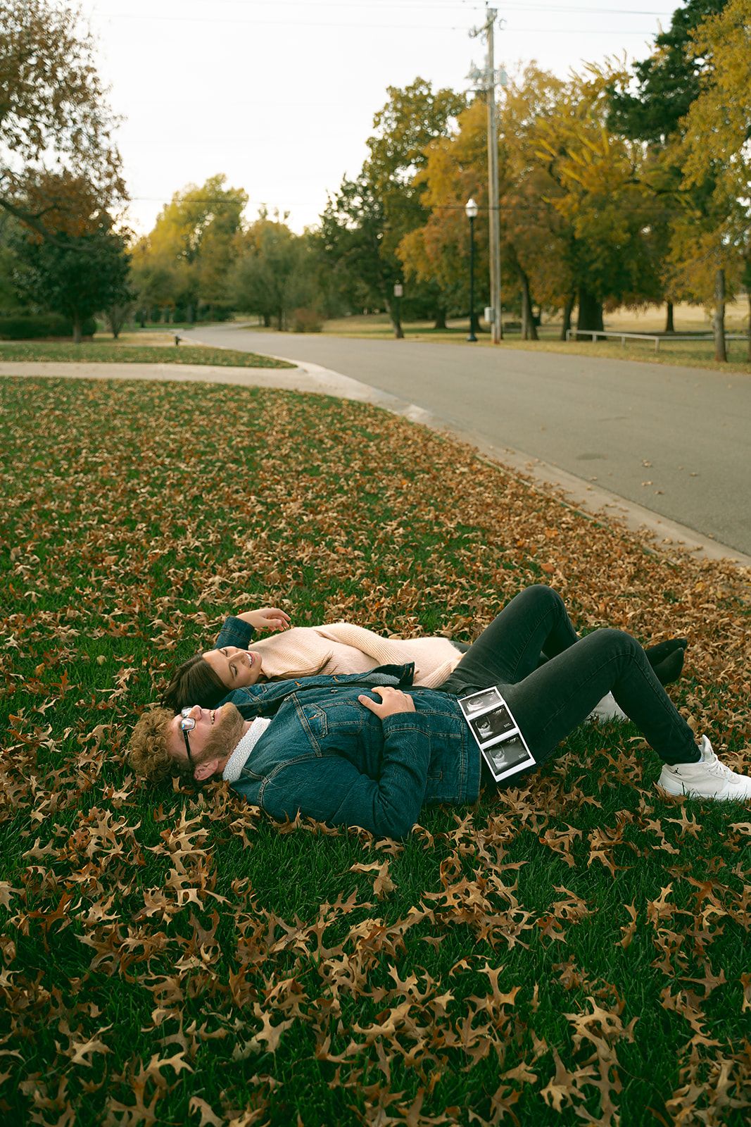 Two people laying on autumn leaves beside paved path in park setting.