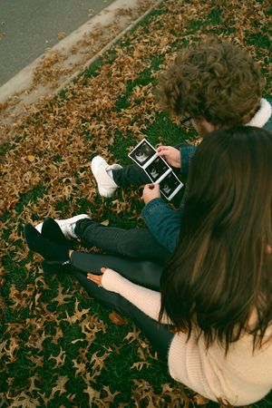 Person in beige sweater and dark pants looking at device while laying on fallen leaves.