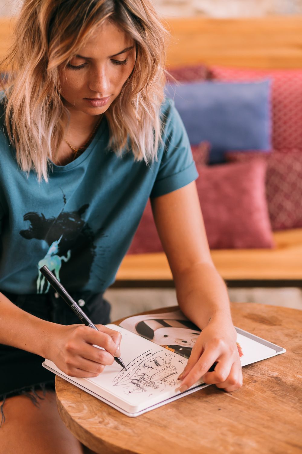 Artist in teal graphic t-shirt sketches in notebook while sitting at wooden table with colorful pillows in background.