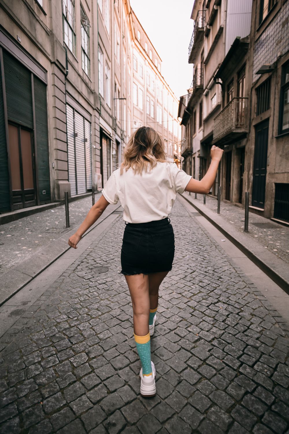 Person in white shirt and black skirt walking down a charming cobblestone street in an old European city.