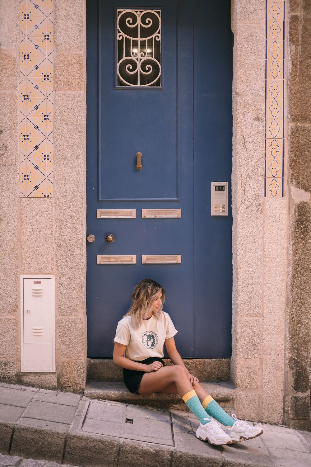 Person sitting on a doorstep in front of a blue door with decorative iron window detail and tiled wall.