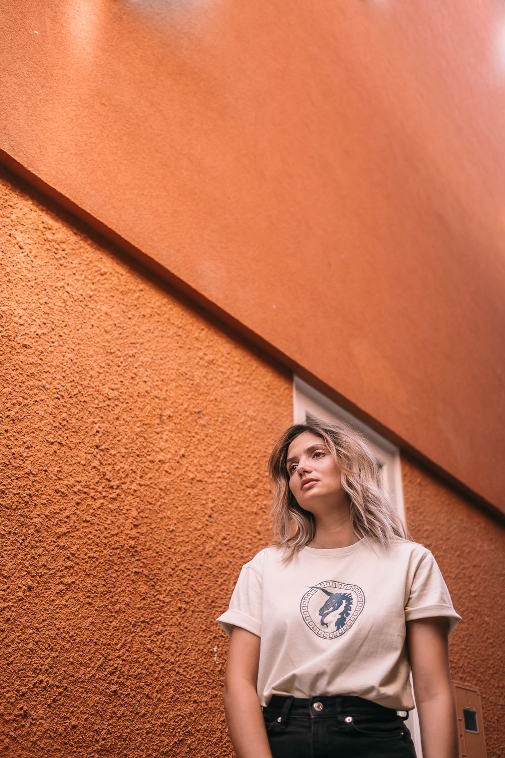 Person wearing a beige graphic t-shirt poses against a textured terracotta-colored wall with diagonal architectural lines.