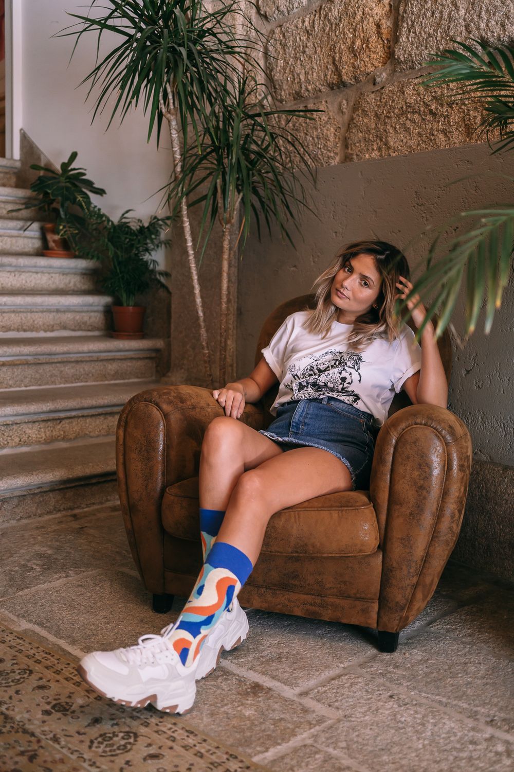 Person relaxing in a brown leather armchair with trendy socks and white sneakers near indoor plants and stairs.