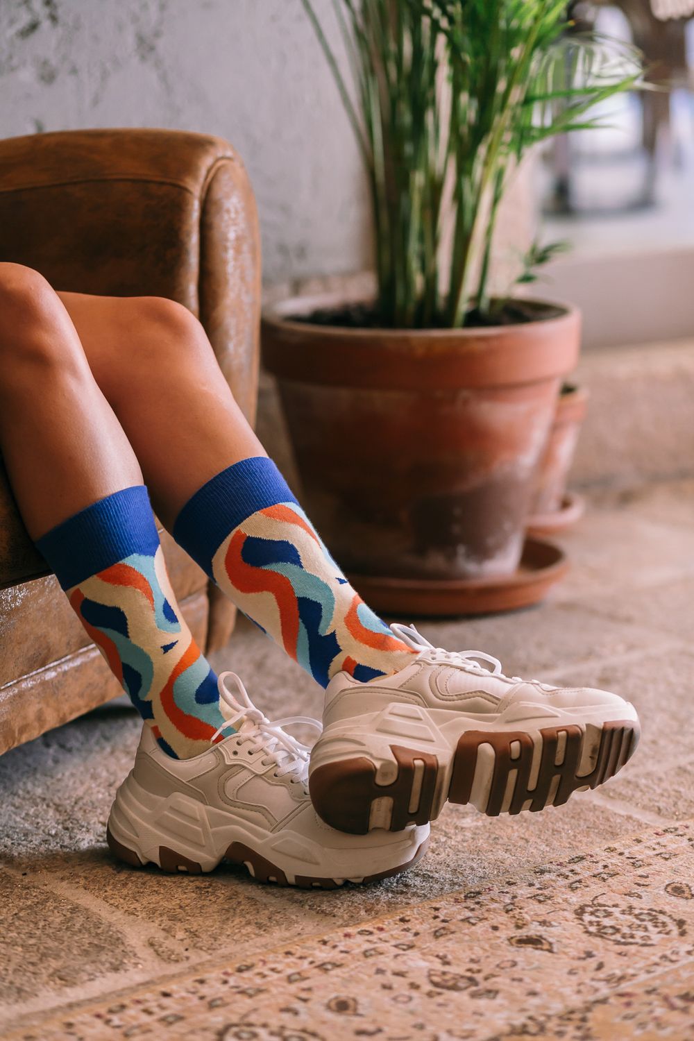 Chunky beige sneakers paired with colorful patterned socks next to a terracotta potted plant on stone floor.