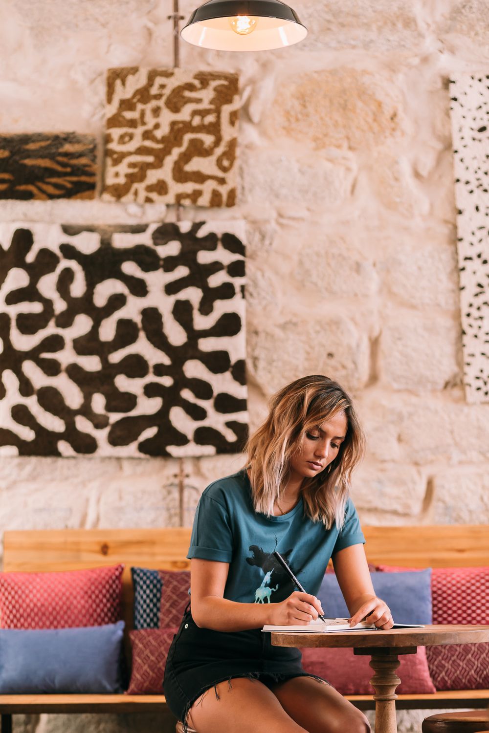 Someone wearing a teal shirt writes in a notebook at a restaurant table beneath abstract wall art and pendant lighting.