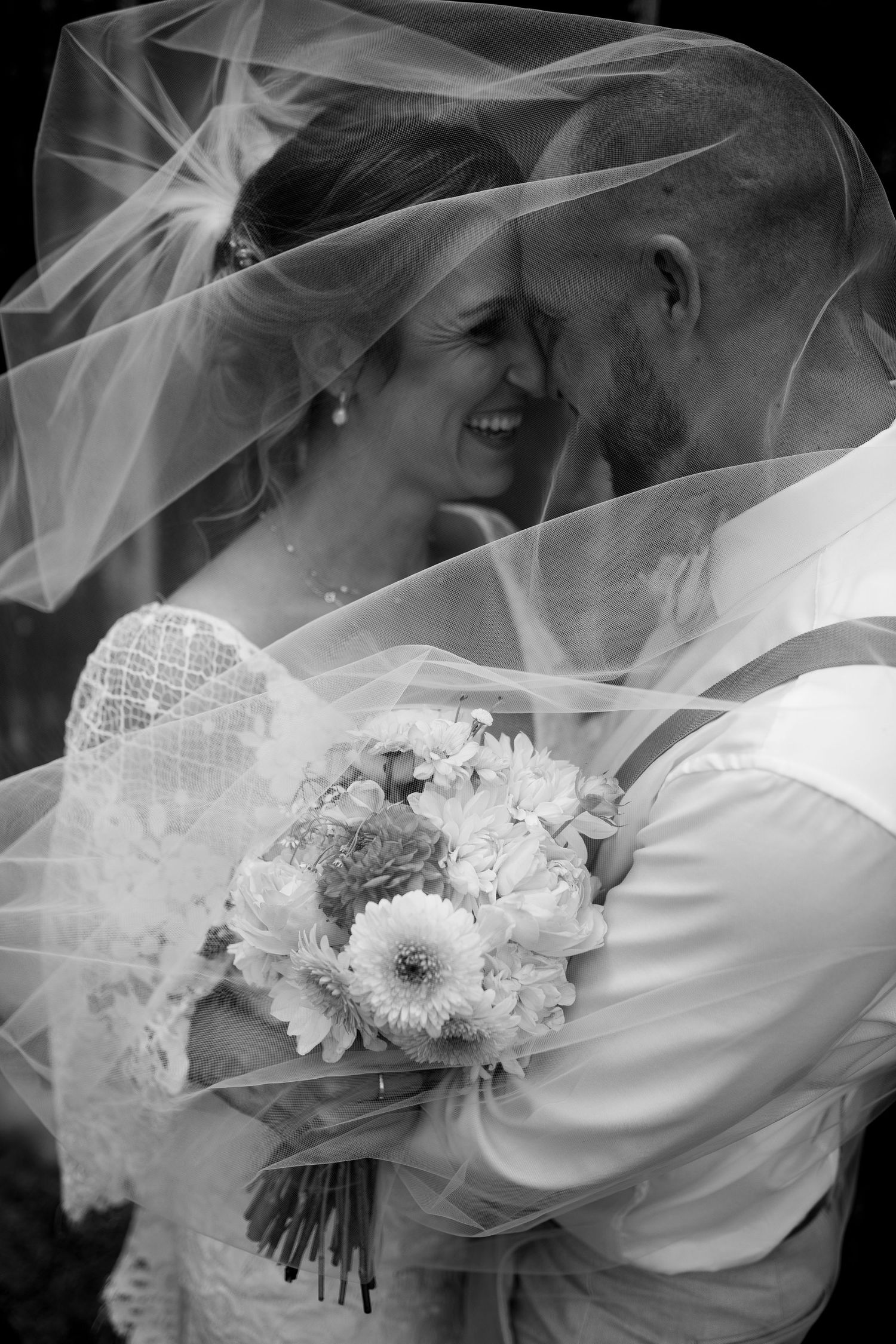 A romantic black and white wedding photo showing a tender moment under a flowing veil with a white floral bouquet.