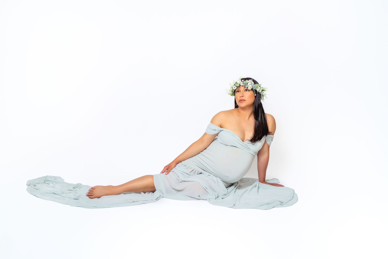 Dreamy maternity portrait with floral crown and flowing pale blue dress on white background.