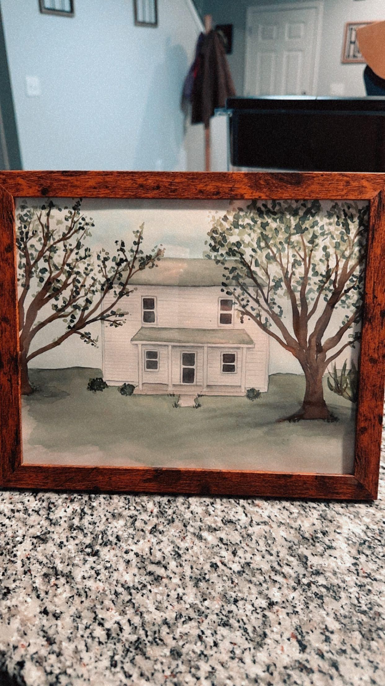 A framed watercolor painting of a white house between two trees on a grassy lawn, displayed on a granite countertop.