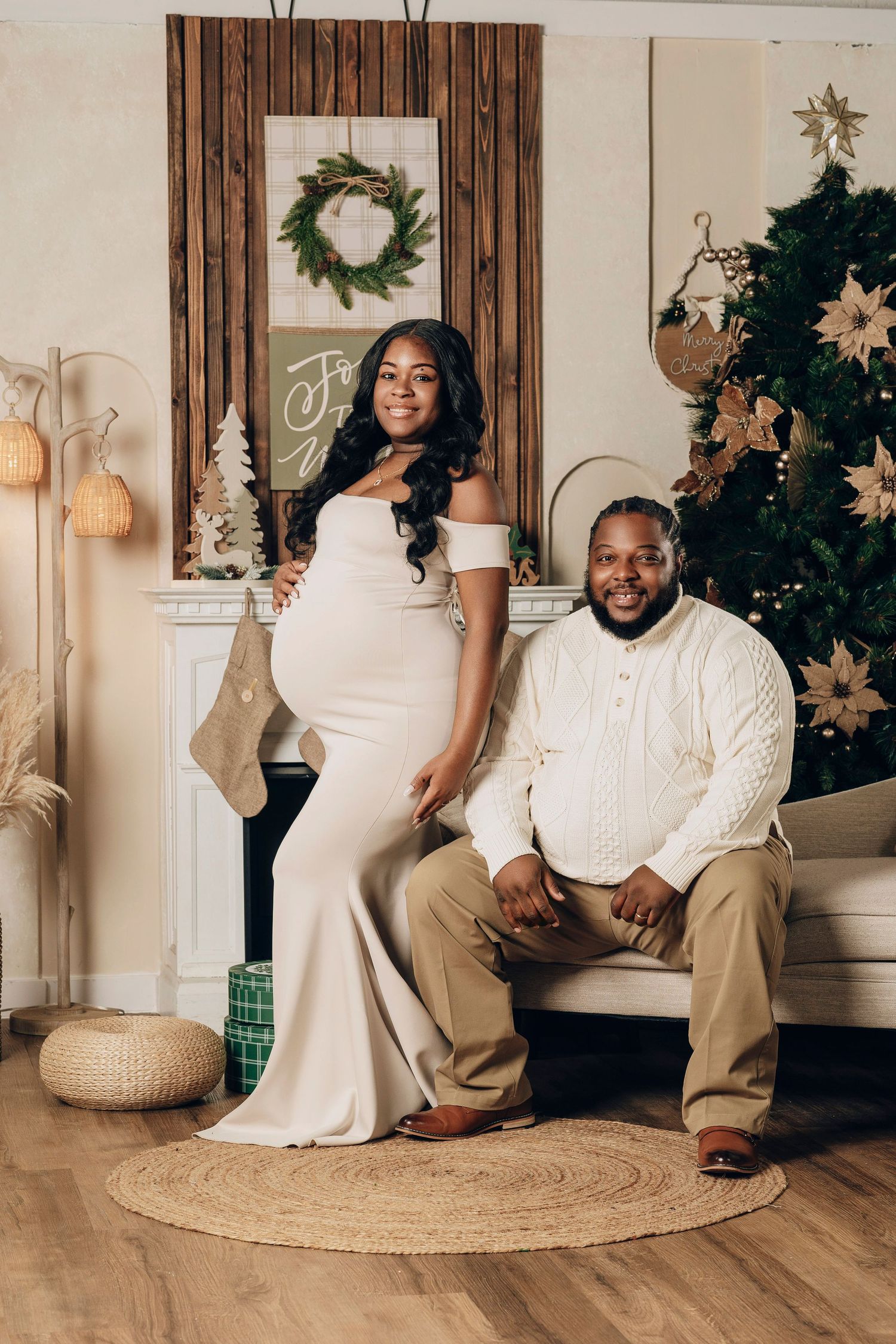 Pregnant woman in white gown poses with partner beside a decorated Christmas tree in a festive holiday setting.