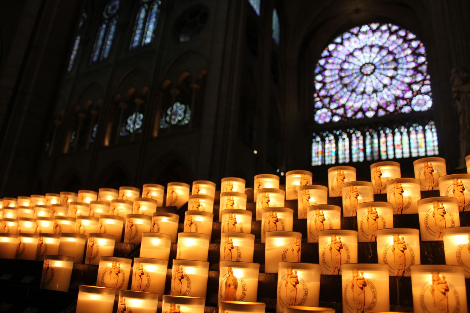 A rose window at Notre Dame cathedral in Pairs with votive prayer candles in front. Dim lighting with vibrant stained glass windows