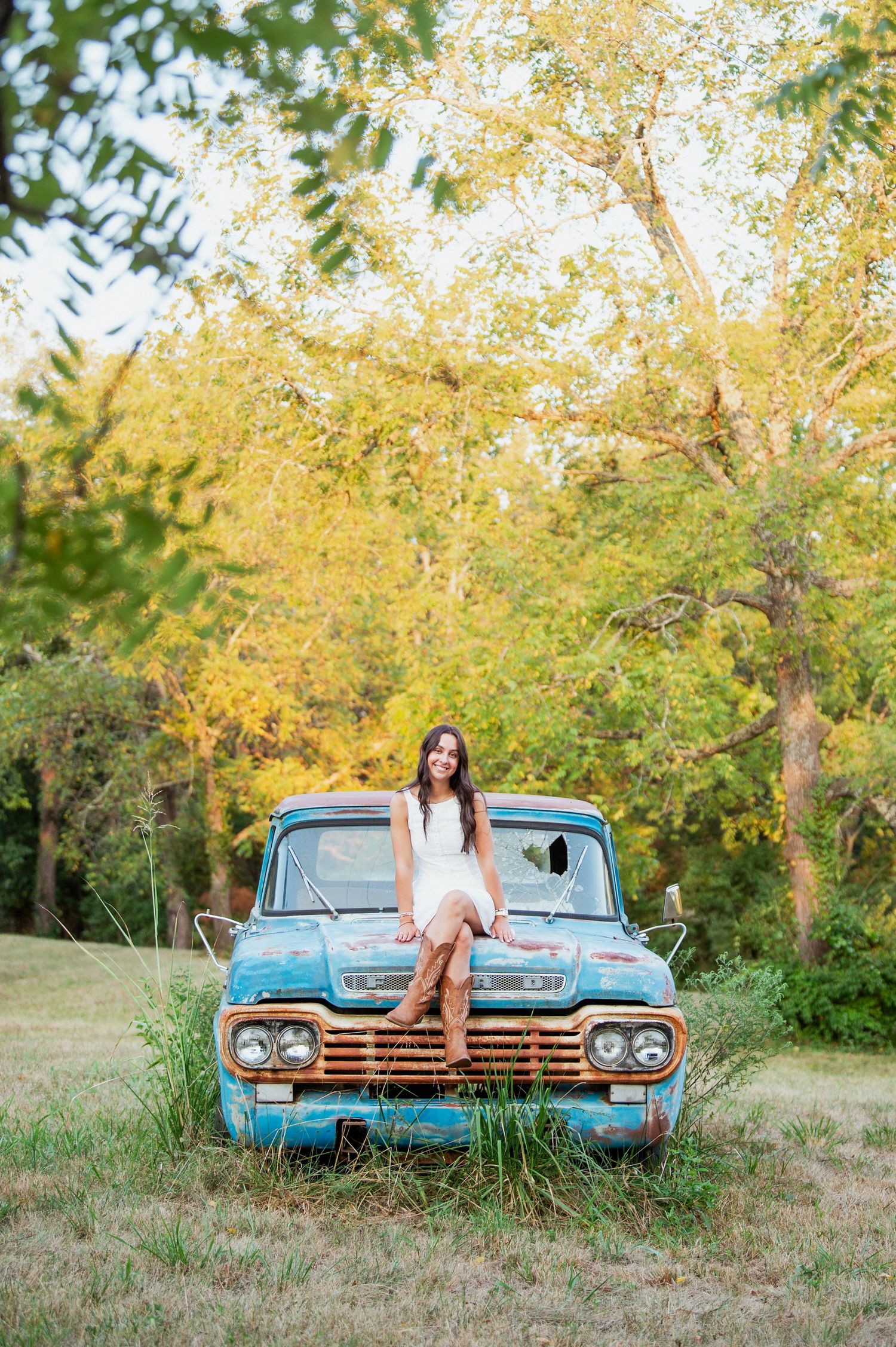 A person in a white dress and cowboy boots reclines on a vintage blue truck in a rural setting.