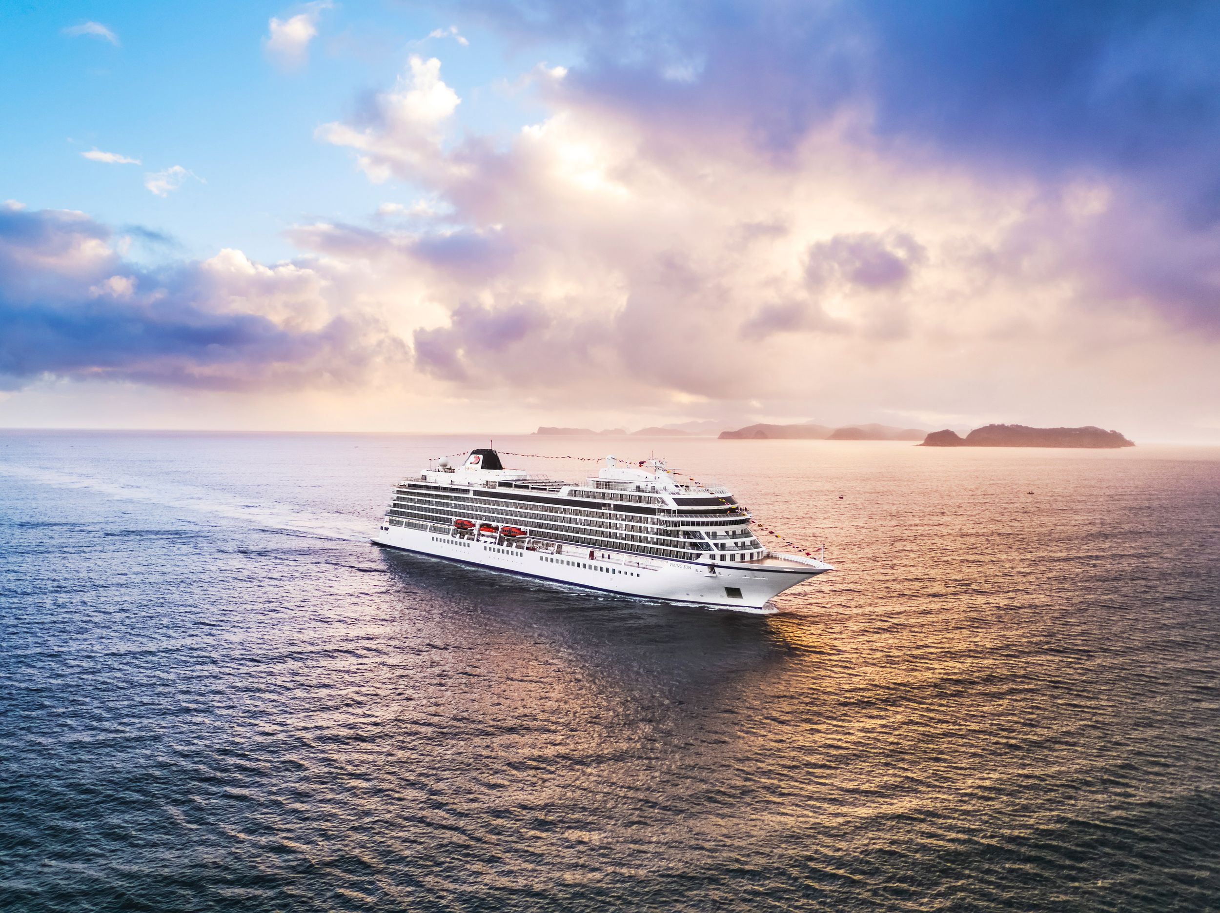 Luxury cruise ship sails across tranquil ocean waters during a vibrant sunset with colorful clouds in the sky.