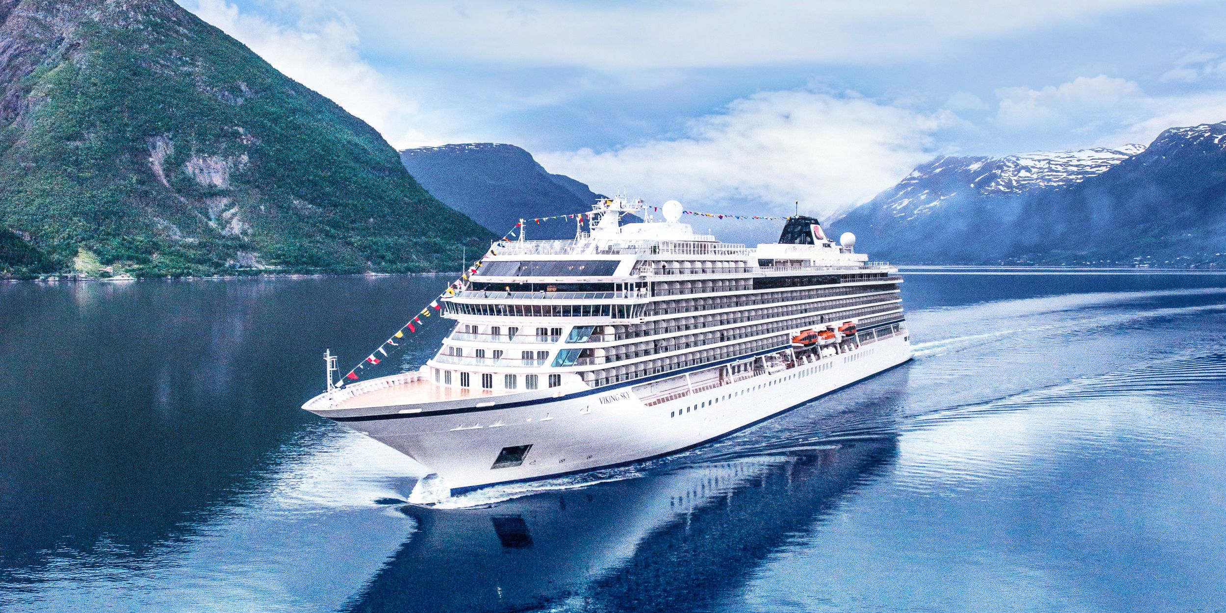 Luxury cruise ship sailing through scenic fjords with snow-capped mountains and blue waters in the background.