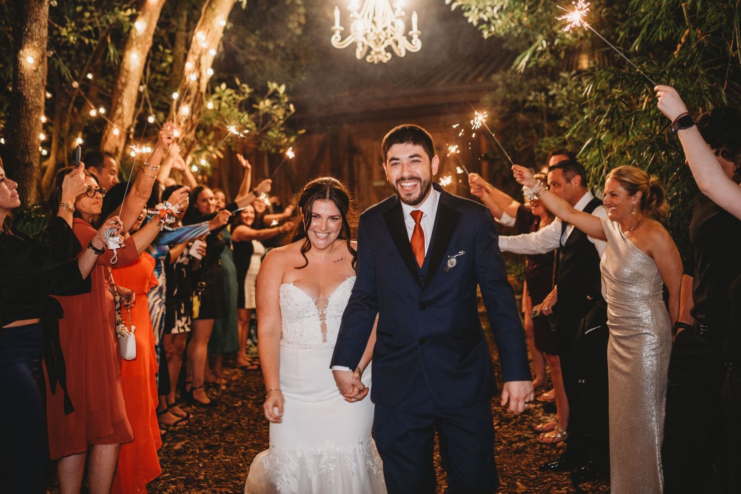 Wedding exit with guests holding sparklers along a tree-lined path at night.