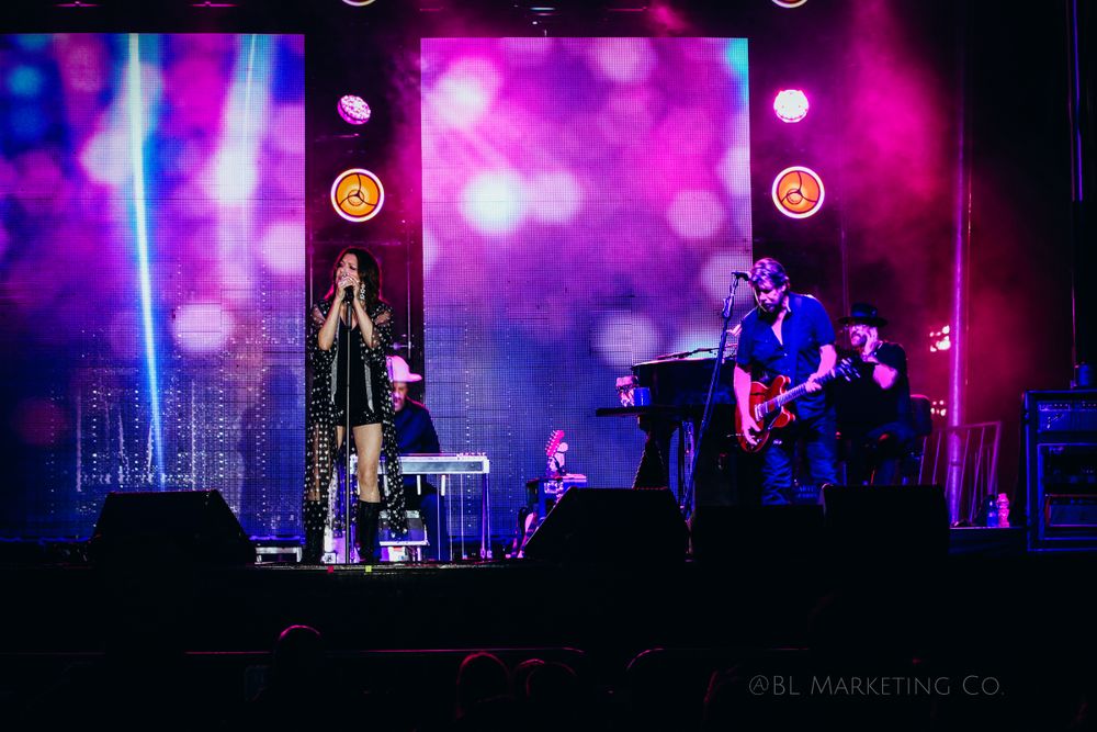 Musicians perform on stage with vibrant purple lighting and visual effects in background.
