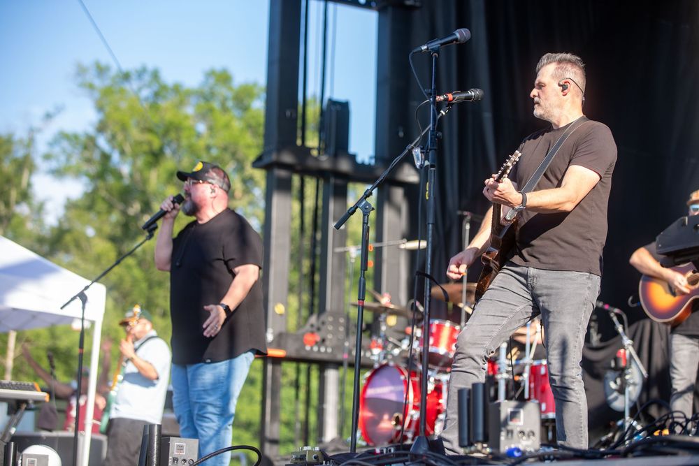 Two musicians perform on outdoor stage, one singing into mic, other playing electric guitar at a live music festival.