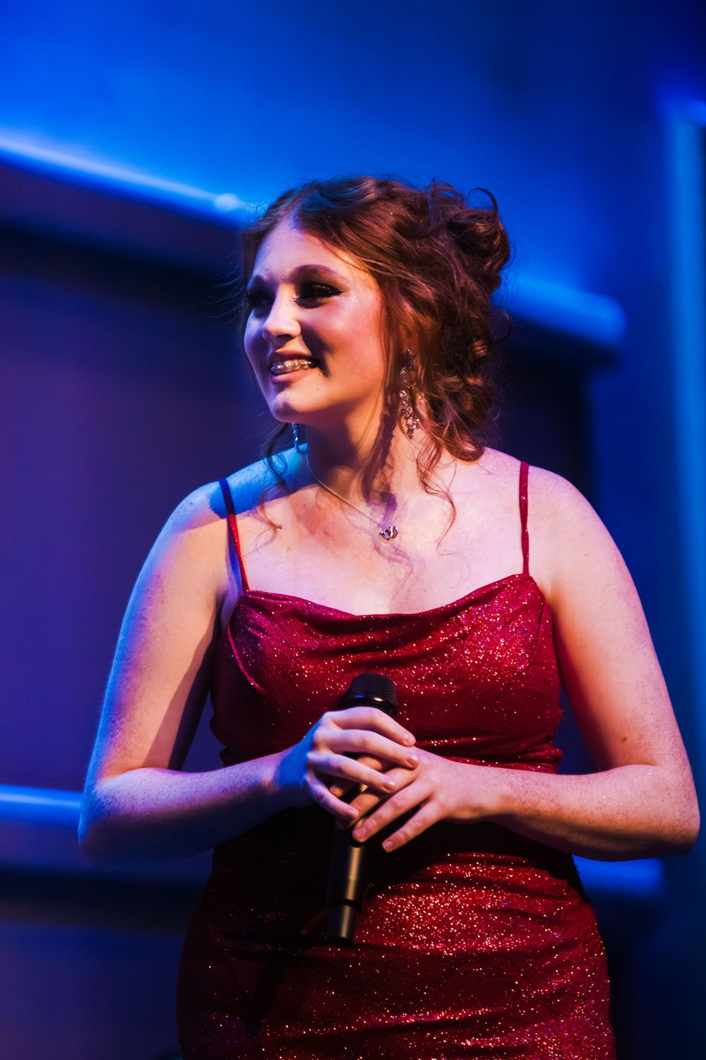 A smiling woman in a red sequin dress holds a microphone under blue and red stage lights.