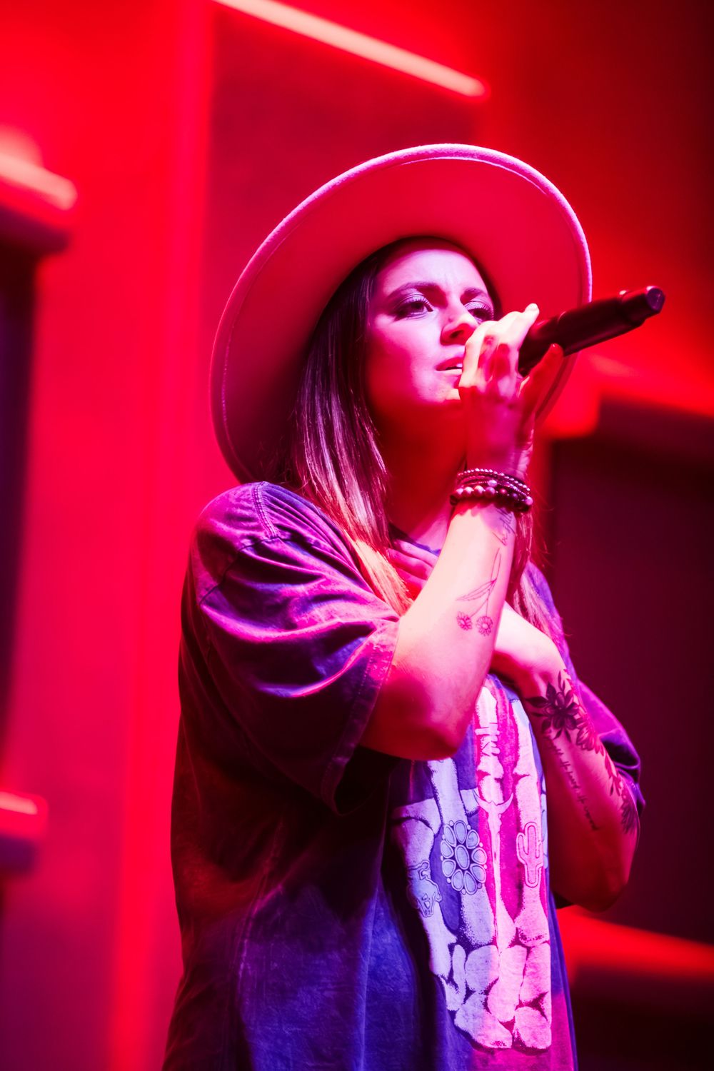 Female singer performing on stage wearing a wide-brim hat, holding microphone under red stage lighting.