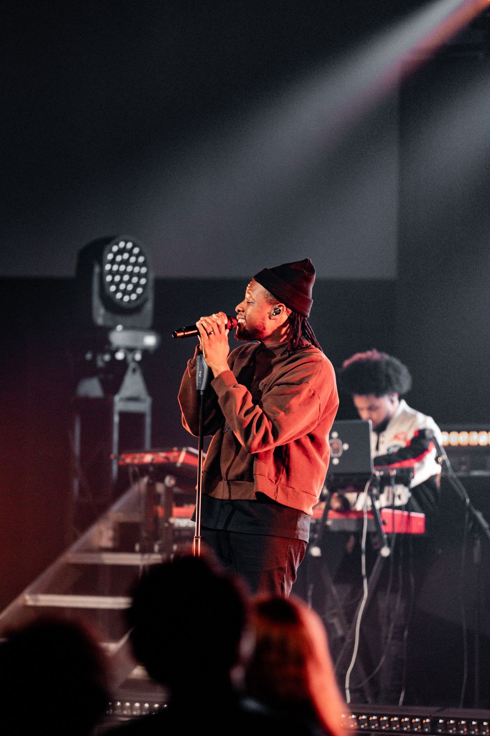 Performer in dark attire on stage with red lighting and band members in background.