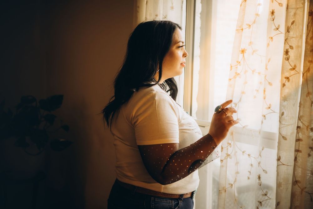 Woman with tattoos gazing out a sunlit window, wearing a white t-shirt in a warmly lit room.