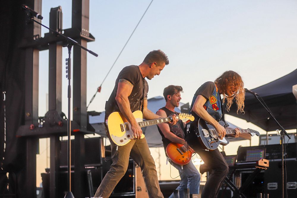 Three guitarists perform energetically on an outdoor stage during a live rock concert at sunset.