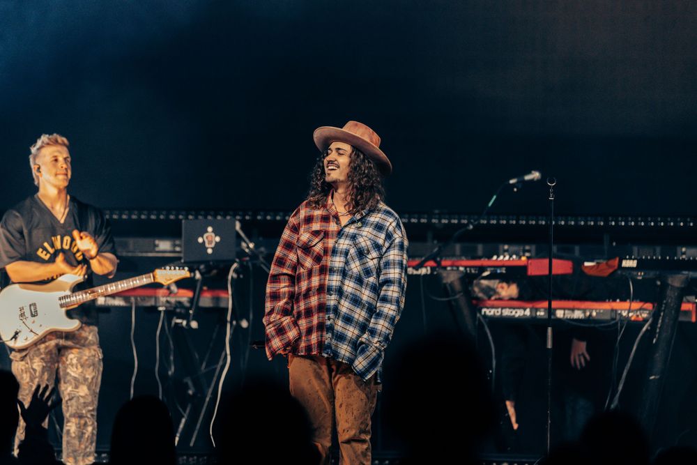 Musicians performing on stage wearing plaid shirts with instruments and stage equipment.