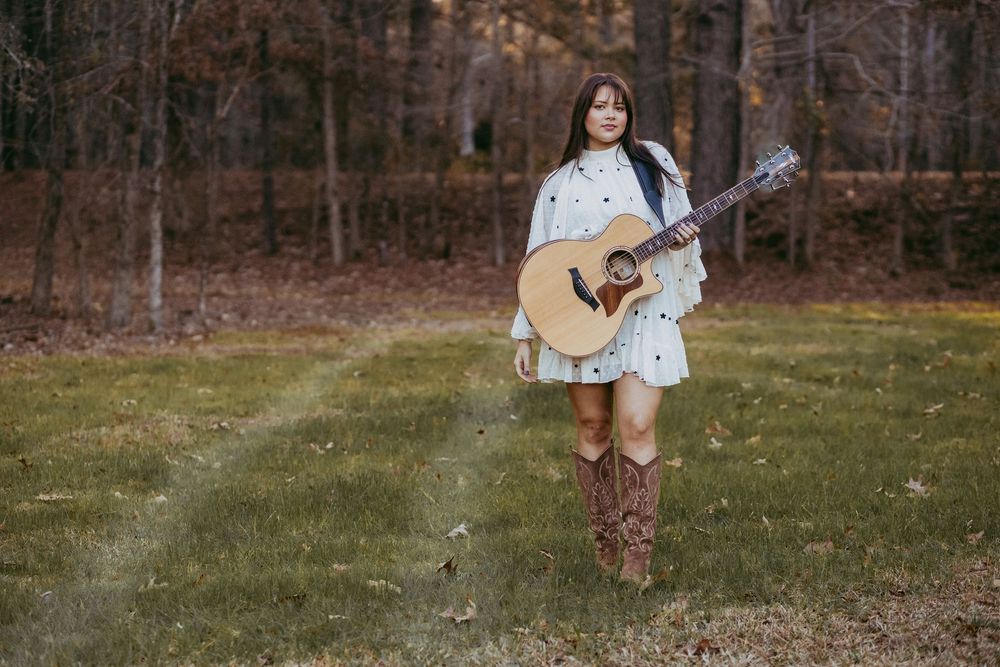 Young woman holding acoustic guitar wearing cowboy boots and white dress, standing on grass with autumn forest background.