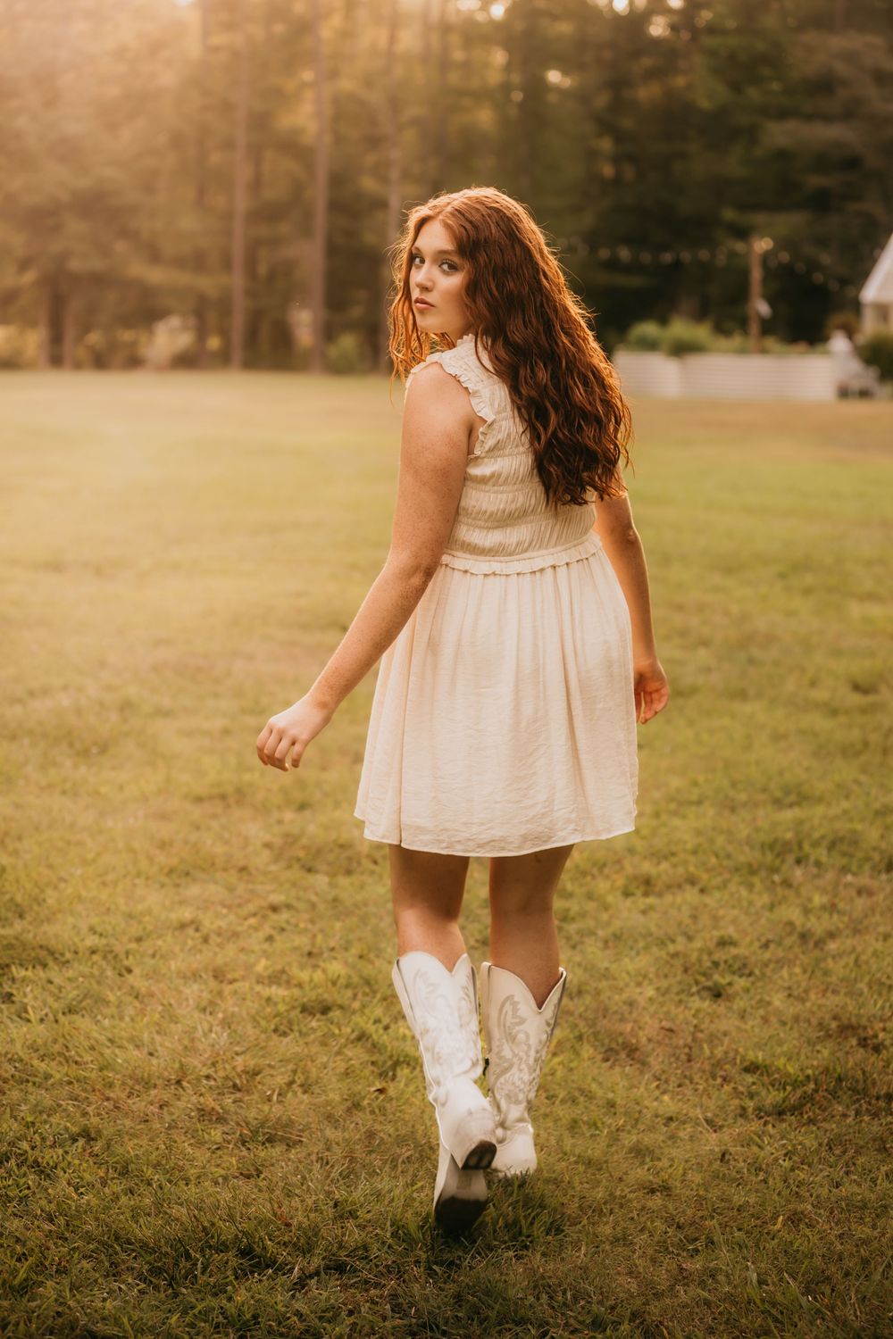 Young woman with long red hair in cream dress and white cowboy boots walking through sunlit grassy field.
