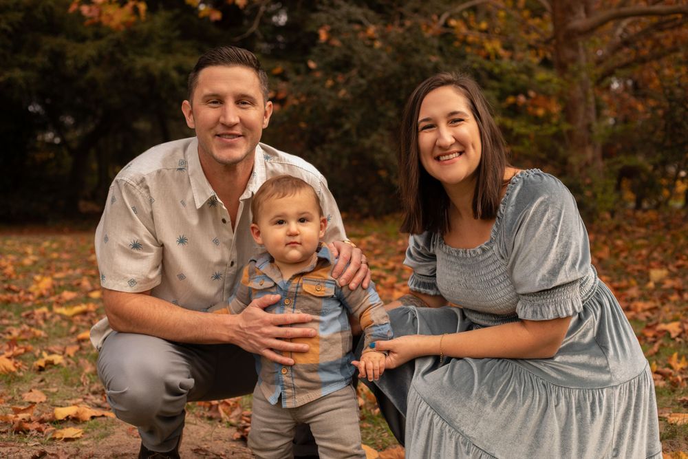 A loving family poses outdoors on autumn leaves with a toddler in blue stripe shirt and gray pants.