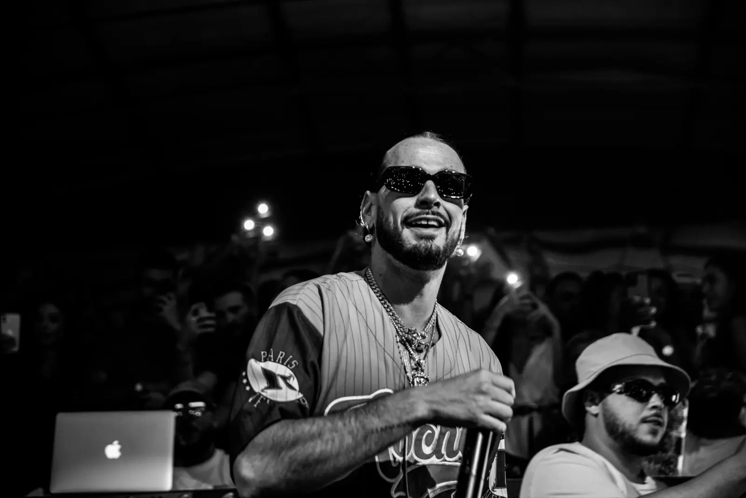 Homme souriant en t-shirt de sport dans une salle de concert, ambiance nocturne avec foule en arrière-plan.