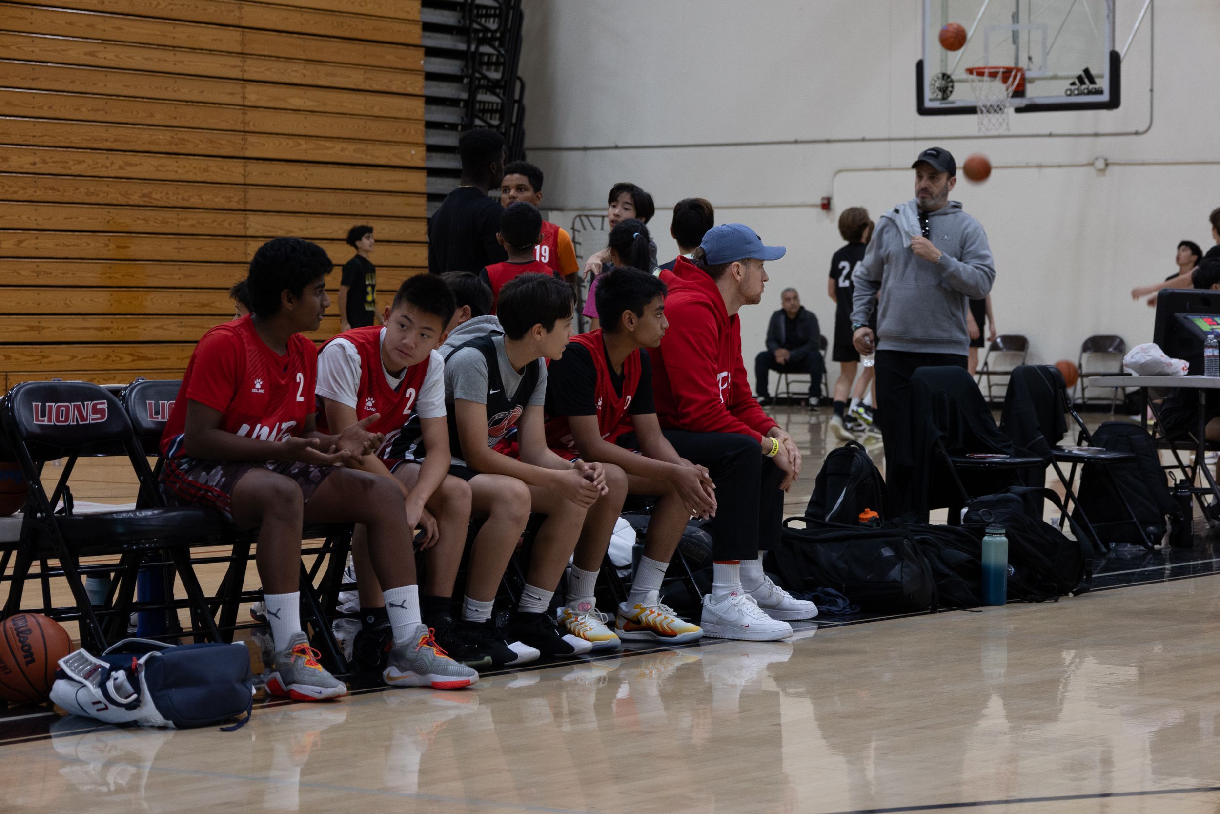 A basketball team sitting on a bench watching their teammates play. Coach is engaged with the game plotting future plans to encourage team