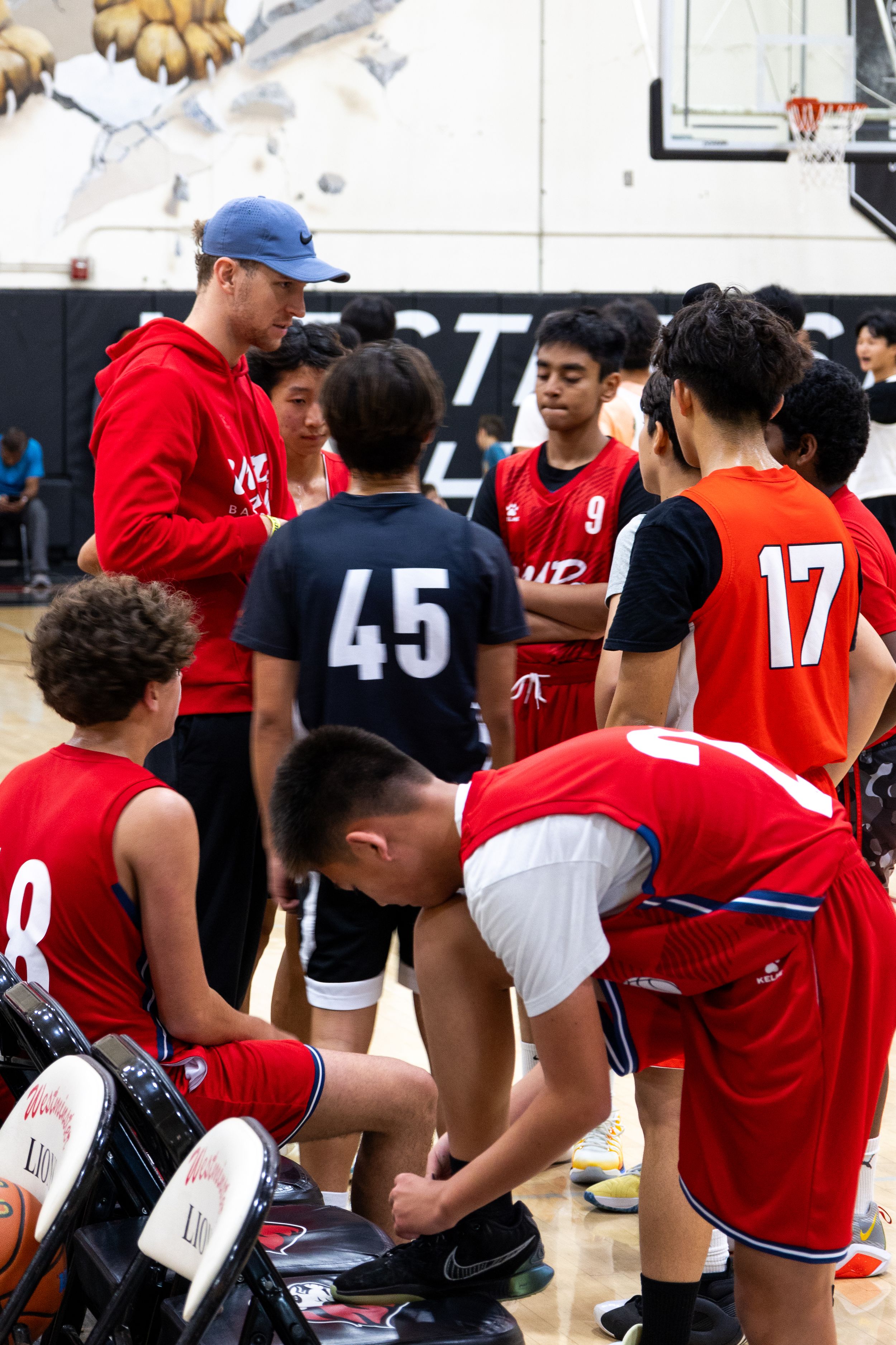A team huddles around the coach while he speak during half time. A player ties his show and another players sits on the bench.