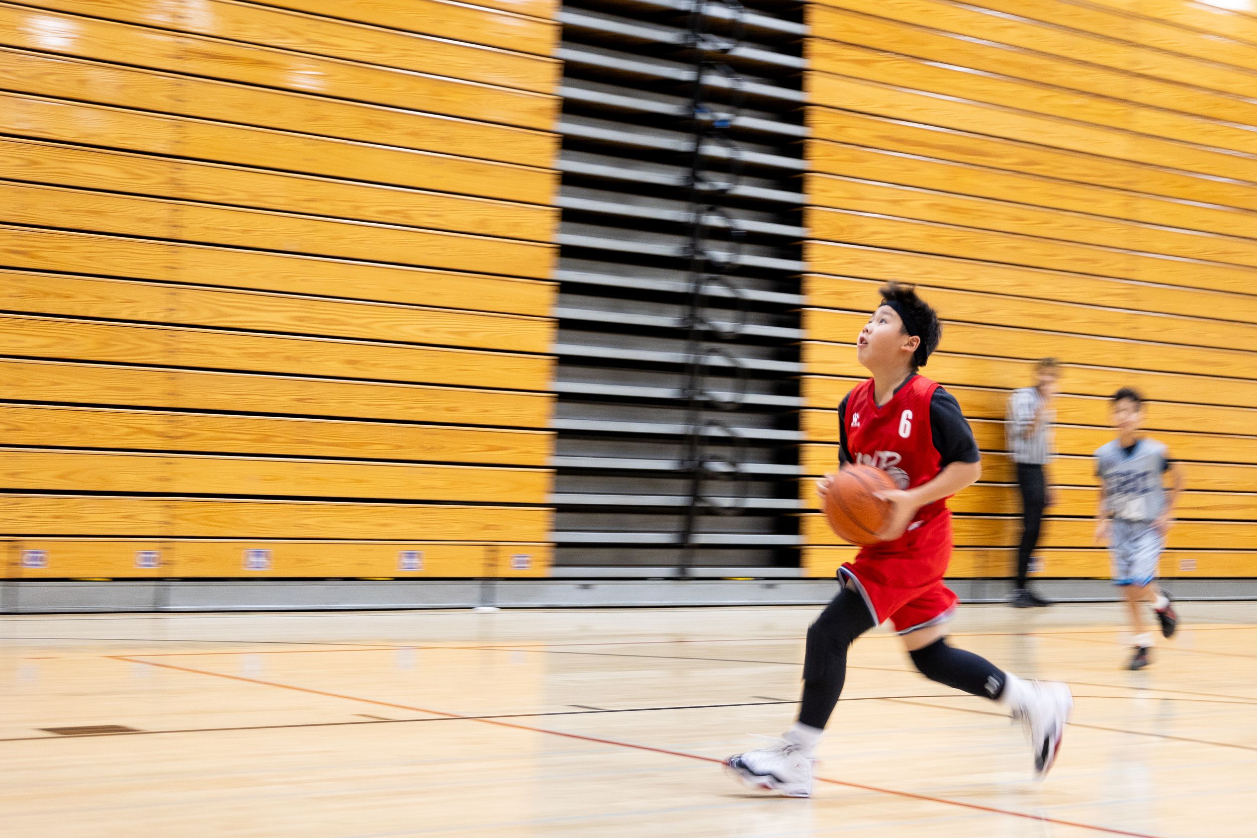 Male youth basketball team player running down the court for a lay-up. The court is in a school gym with bleachers in the background.