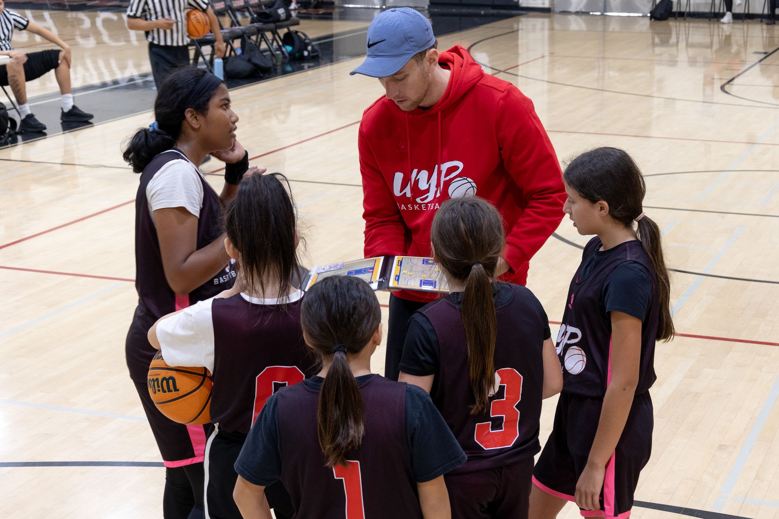 Candid photography of a basketball coach leading a team huddle during a time out showing game plans on a clip board and listening to players