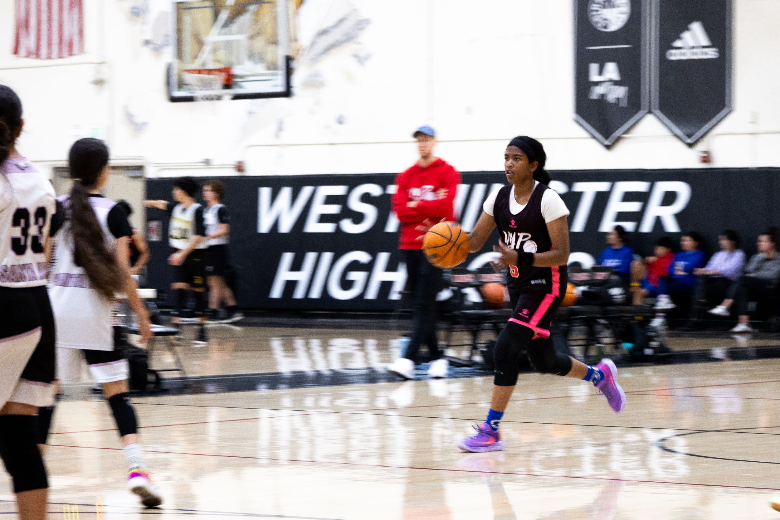 An action photograph of a female basketball player running and dribbling a ball while the background of her coach, team and court are blurry