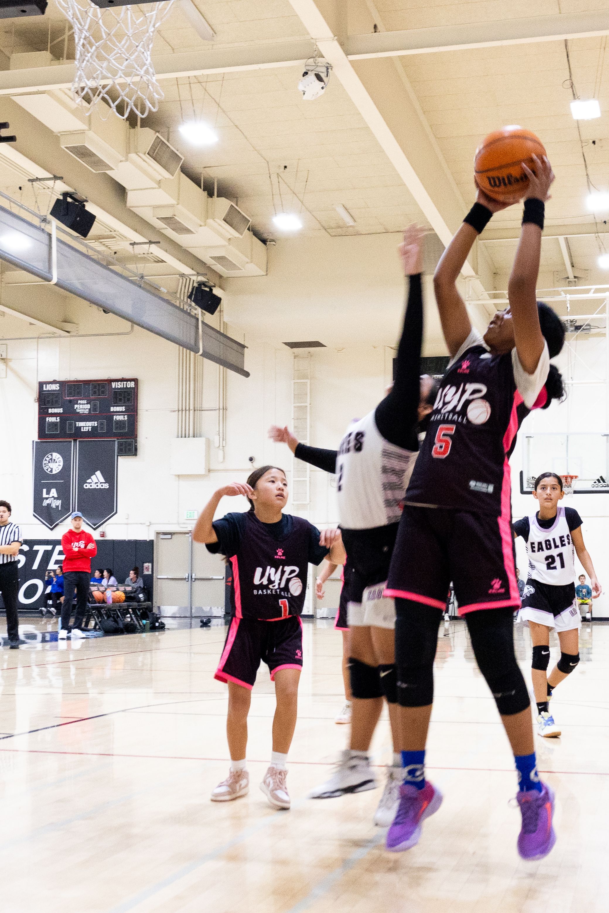 A candid action photo of a female youth basketball player midjump shooting a basketball into a hoop while a defender is trying to block