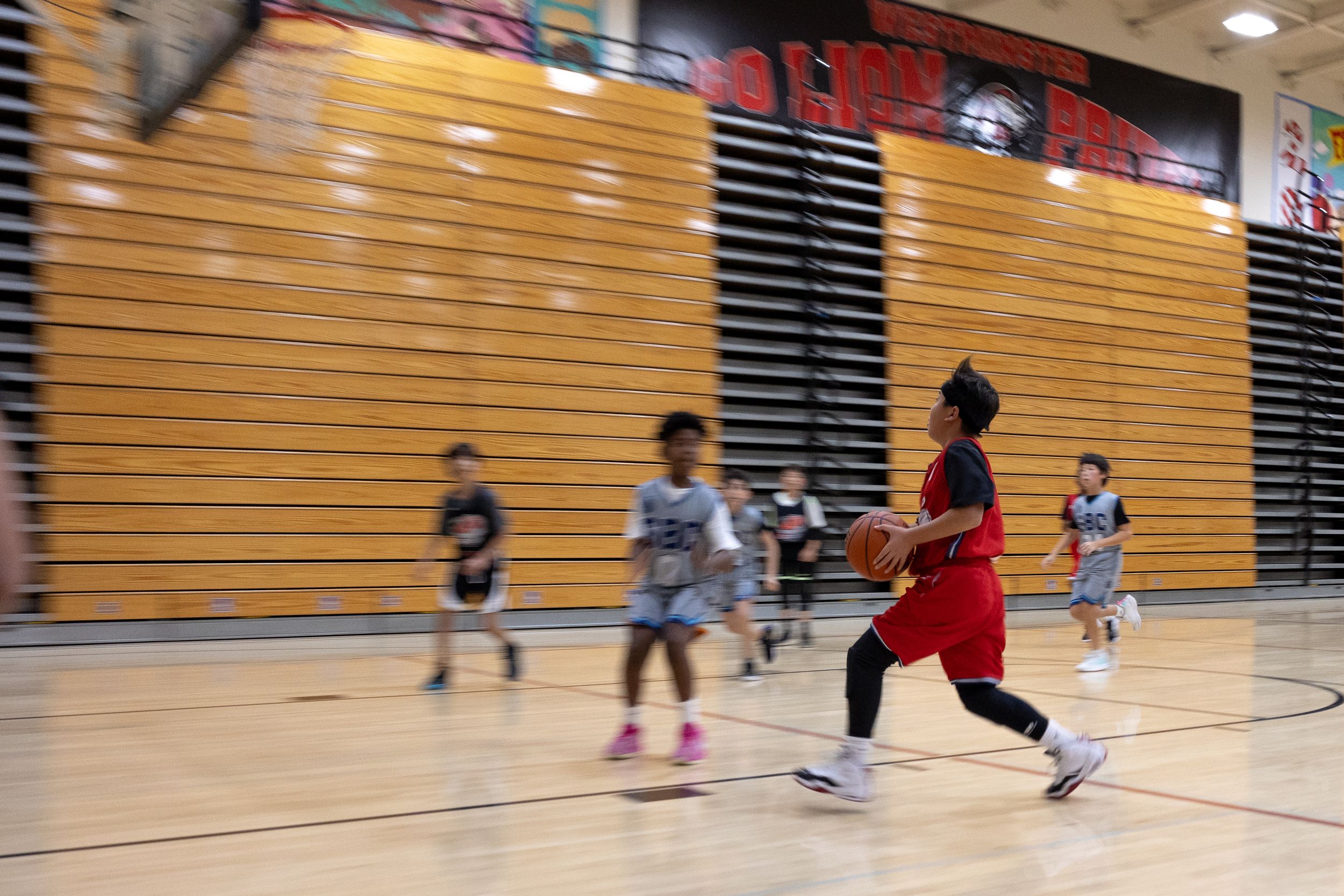 An action photograph capturing a youth basketball player running to make a layup during a game. Background is blurry with the player focused