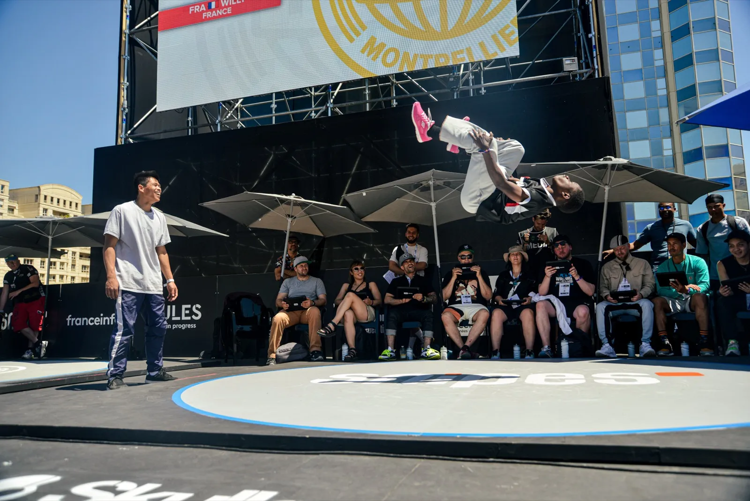 Figure de breakdance en plein saut sur une scène, sous les regards d’un jury et du public.