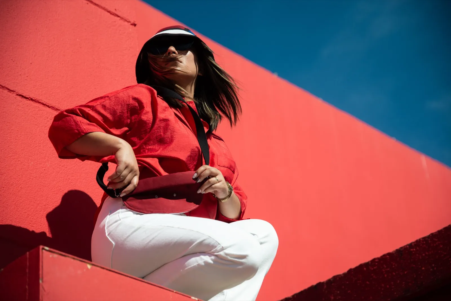 Femme posant contre un mur rouge intense sous un ciel bleu, vêtue d’une veste rouge et d’un pantalon blanc, regard vers le haut.