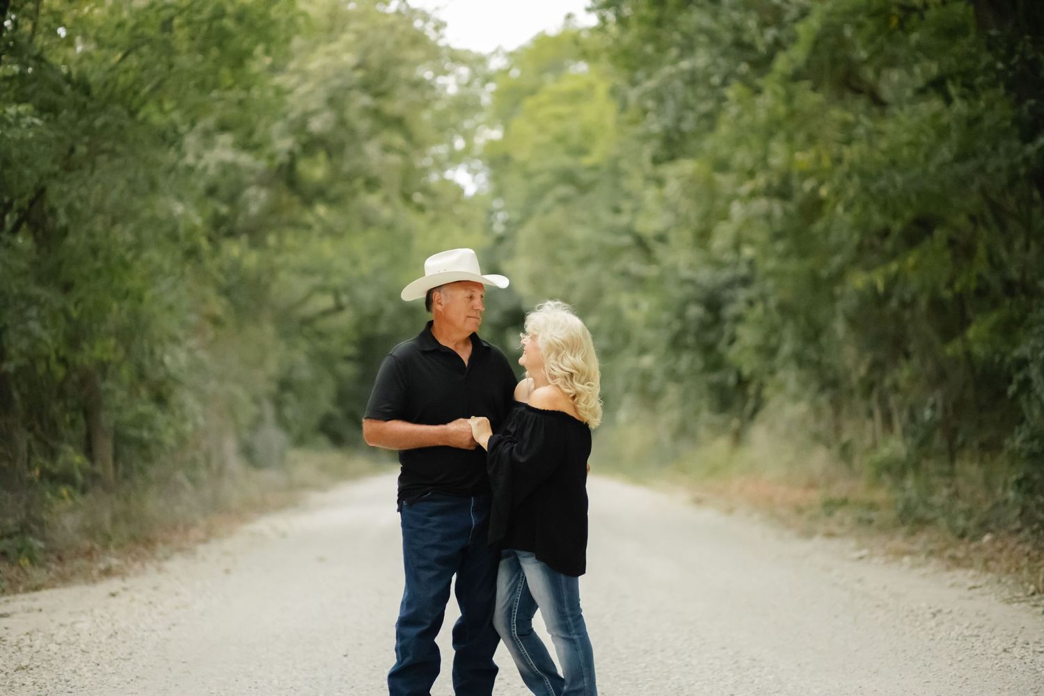 A couple in western wear dancing together on a tree-lined gravel road in a romantic outdoor setting.