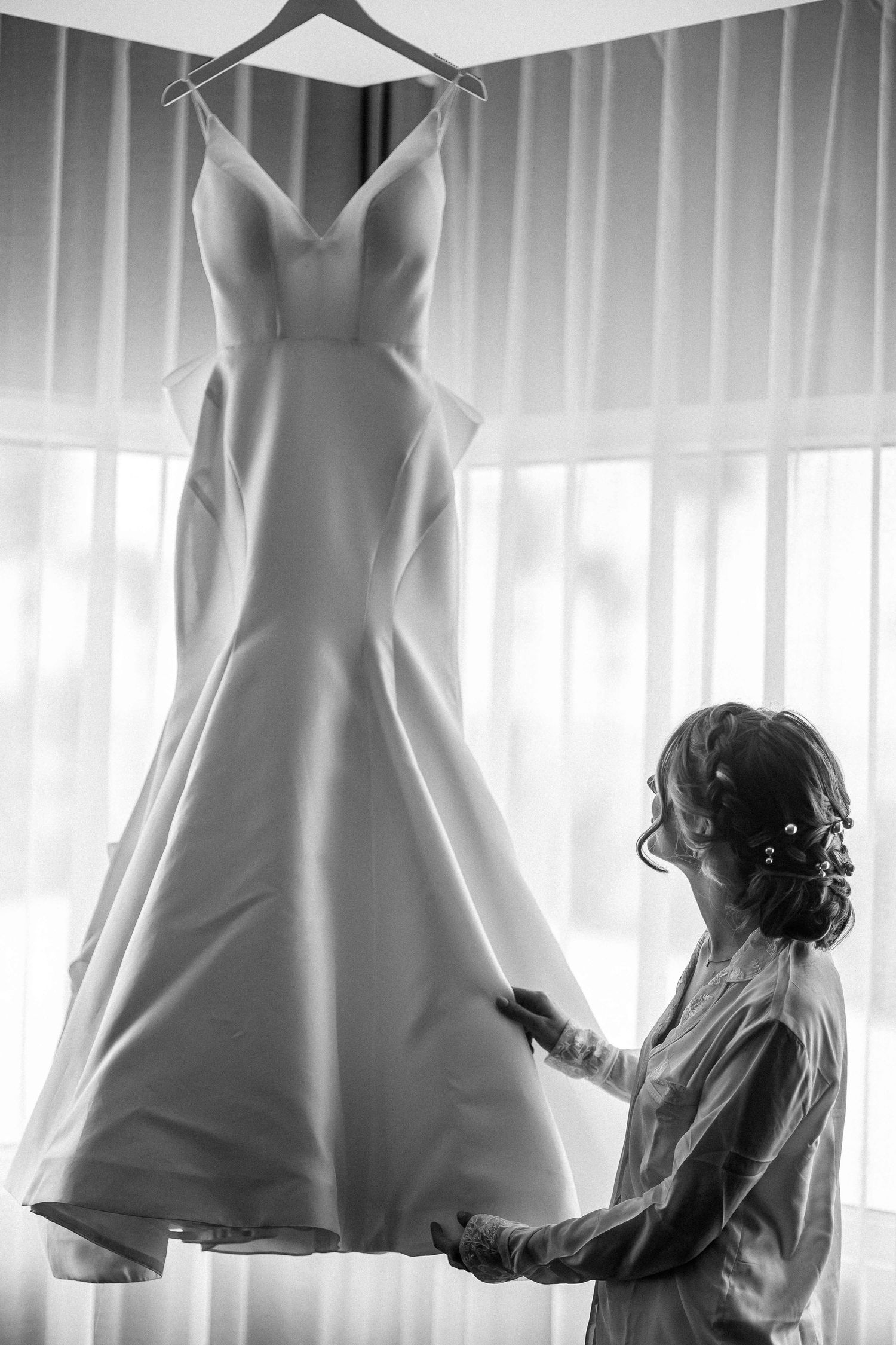 Classic white wedding dress displayed on hanger against sheer curtains.