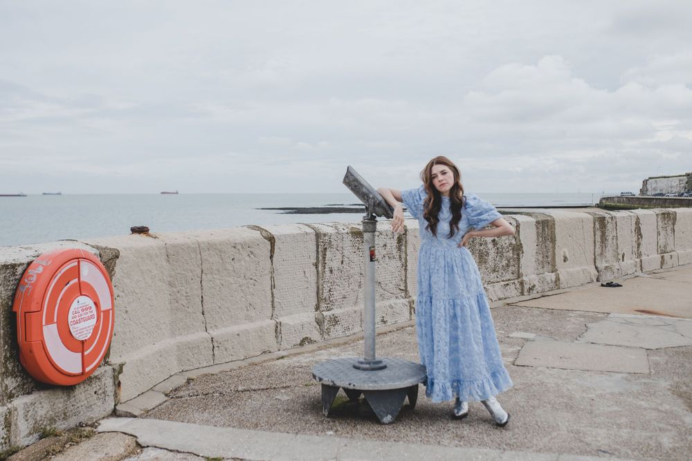 Woman in flowing blue dress posing dramatically by coastal viewfinder.