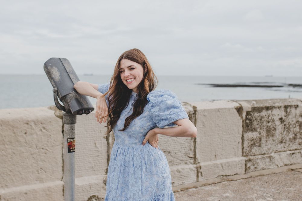 Person in vintage blue dress poses playfully by seaside viewing station.