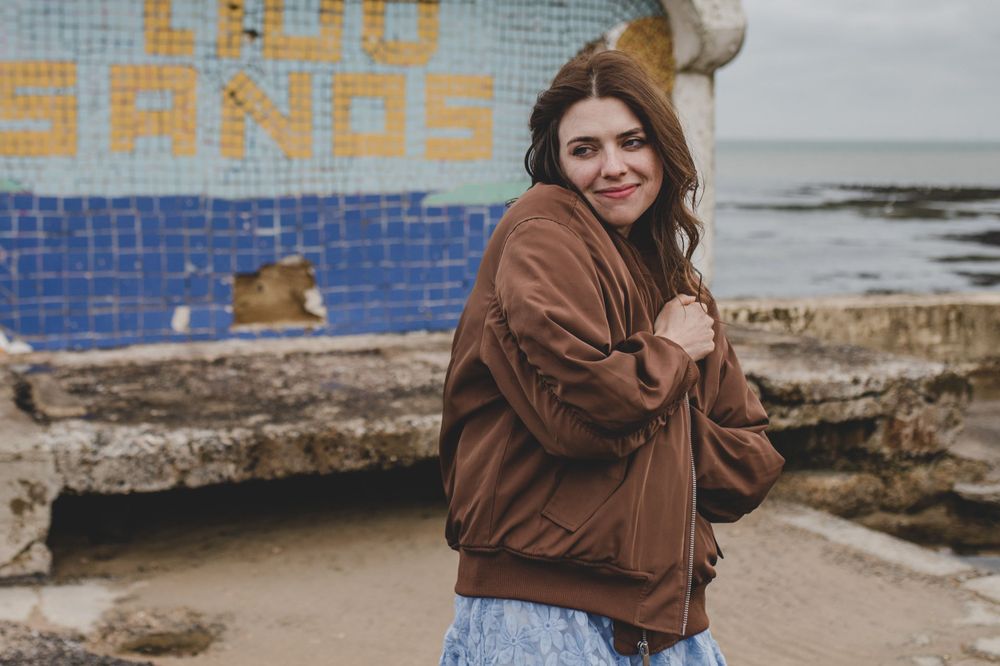 Person in brown jacket poses playfully near blue wall at beach.